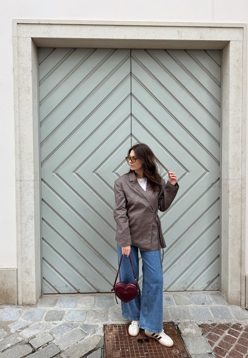 Blazer marron ceinturé, chemise blanche, jean large bleu, sac bordeaux en forme de cœur, baskets blanches, debout devant une porte chevron vert clair.