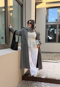 Manteau en laine gris, haut péplum gris clair, pantalon large blanc, sac à main noir. Lunettes de soleil foncées, écharpe superposée ; sol enneigé ; toile de fond d'architecture moderne.
