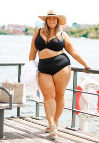 Černý bikiny set s podpůrným topem a vysokými kalhotkami. Modelka má na sobě široký slamáky a sandály na šněrování. Kousek vedle je neutrální kabelka.