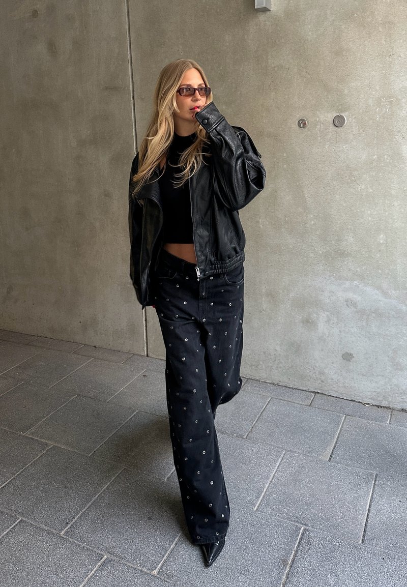 Schwarze Leder-Bikerjacke, eng anliegendes schwarzes Crop-Top, schwarze Hosen mit dekorativen silbernen Ösen, spitze schwarze Absätze, die vor einem Beton-Hintergrund gehen.
