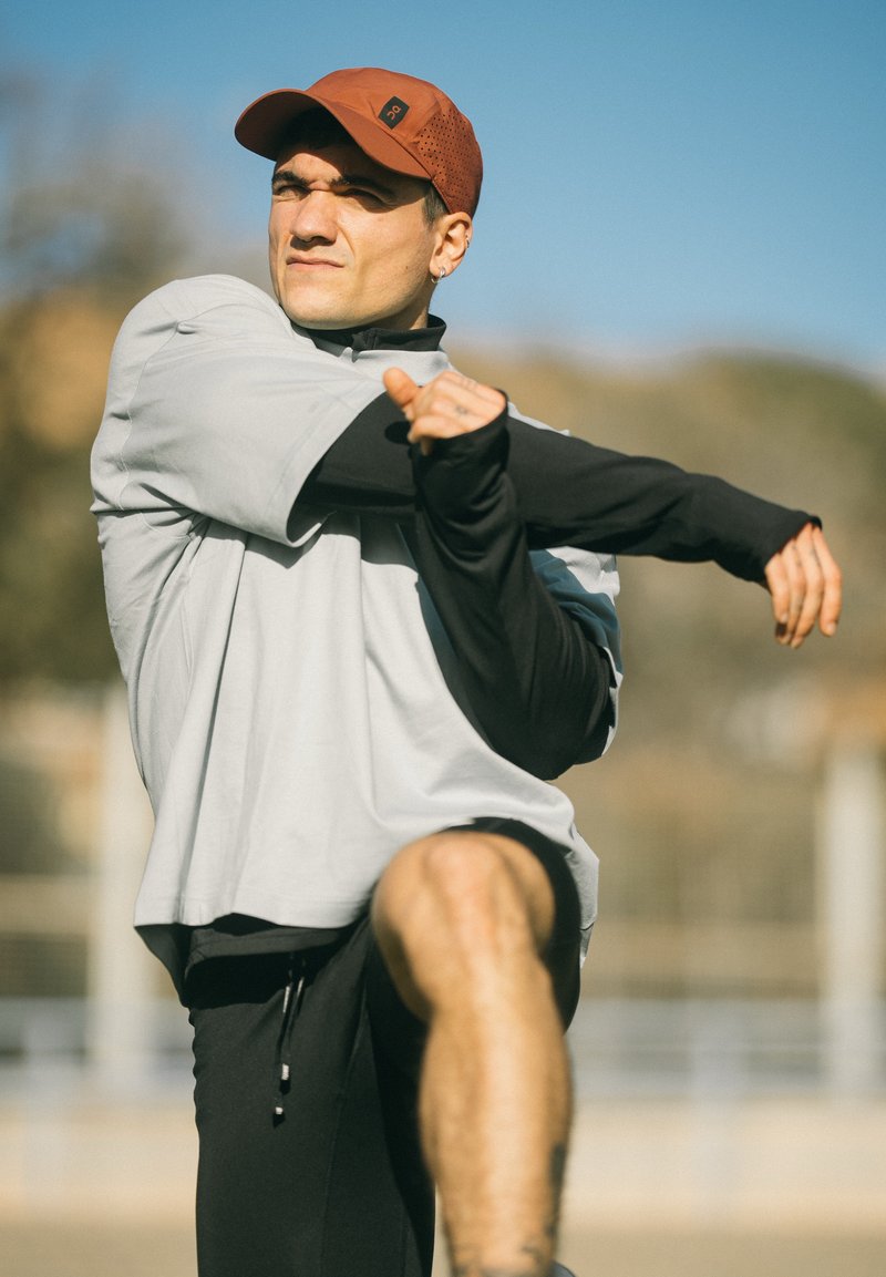 Man met een rode pet en sportkleding die zijn arm over zijn borst uitstrekt, buiten op een zonnige dag.