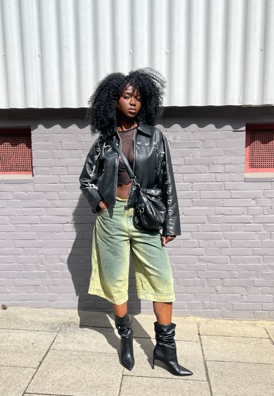 Chaqueta de cuero negra cropped con ojales plateados, blusa transparente, vaqueros holgados en tonos verde amarillento y botas negras de tacón hasta la rodilla.