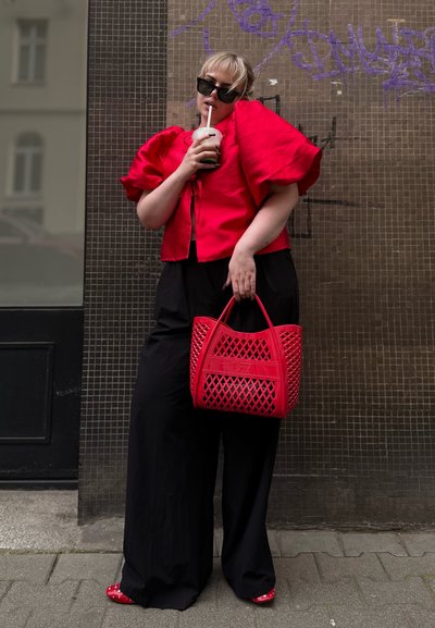 Top rojo de mangas abullonadas, pantalones de pierna ancha negros, bolso perforado rojo y zapatos rojos con lunares blancos. El fondo presenta una pared con un patrón.