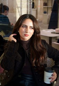 Mujer con un abrigo oscuro texturizado y un cuello alto, sosteniendo una taza de café, cabello largo y ondulado, maquillaje sutil, sentada en un ambiente de cafetería.