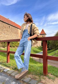 Ανοιχτό μπεζ πουκάμισο επάνω από ένα λευκό τοπ, μπλε τζιν σαλοπέτα, γκρι κάλτσες και καφέ κοκάλια. Εξωτερικός χώρος με πέτρινο τοίχο και πράσινα φυτά.