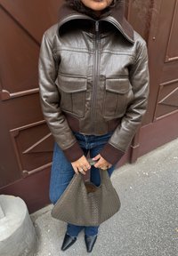 Brūna ādas saīsināta jaka ar ribotu brūnu apkakli, divām priekšējām kabatām, saskaņota ar ziliem džinsiem un austu pelēku rokassomu. Norādīti melni apavi.