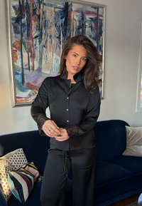 Femme aux longs cheveux bruns portant un vêtement de détente en satin noir avec des poignets en dentelle, debout devant un canapé bleu et une peinture abstraite.