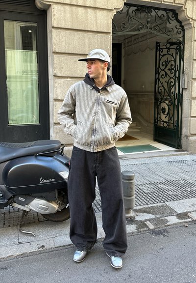 Chaqueta gris a rayas con cremallera sobre una sudadera negra, combinada con pantalones anchos negros y zapatillas plateadas brillantes. Un patinete negro está aparcado cerca.