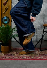 Personne retroussant le pantalon pour montrer des bottes de randonnée marron, debout sur un tapis à motifs près d’un fougère en pot et d’un meuble en bois avec une décoration musicale.