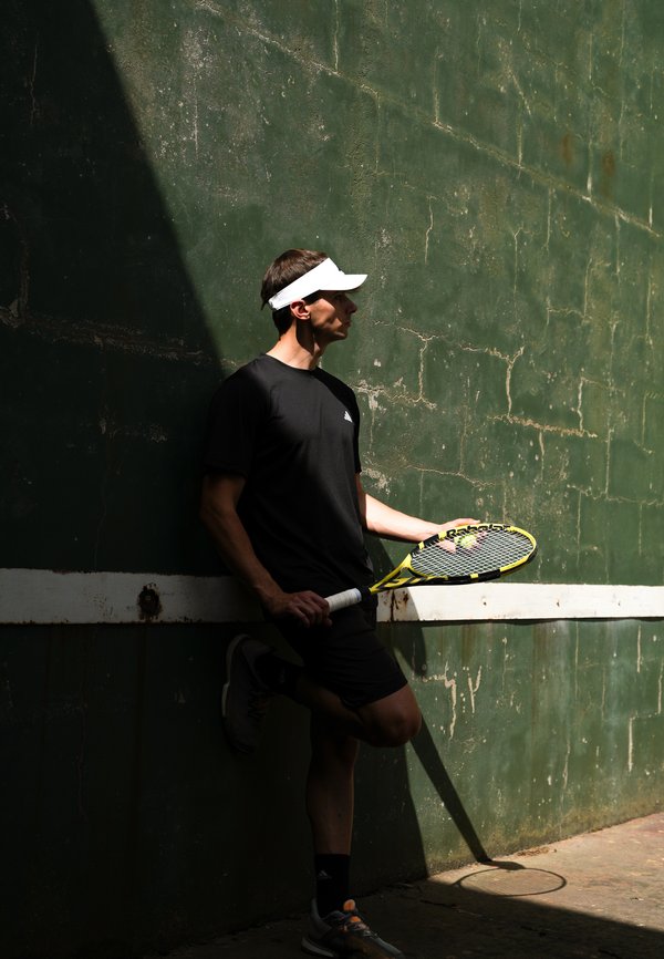 Man in zwarte sportkleding en wit vizier die een tennisracket vasthoudt, leunend tegen een groene muur in gedeeltelijk zonlicht en schaduw.