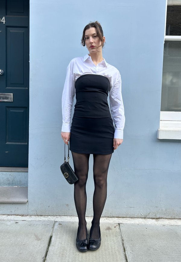 Donna in piedi contro una parete azzurro chiaro, indossa un vestito nero senza spalline sopra una camicia bianca con bottoni, collant neri e ballerine nere, tenendo una piccola borsa nera.