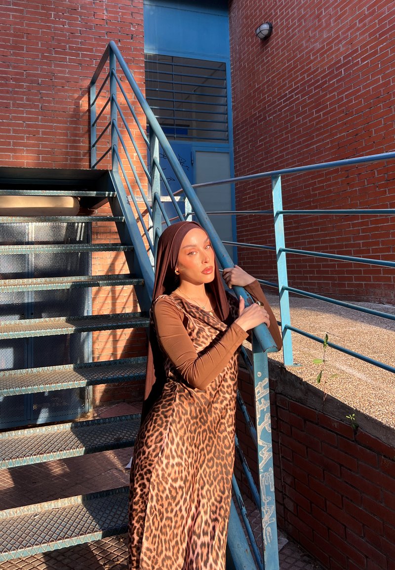 Femme portant un hijab marron et une robe à imprimé léopard appuyée sur une rambarde métallique bleue près d'un escalier extérieur avec des murs en briques rouges au soleil.