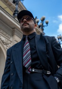 Completo blu a doppio petto, camicia nera e una cravatta a righe rossa e grigia. Gli accessori includono occhiali da sole e una cintura nera con fibbia argentata.