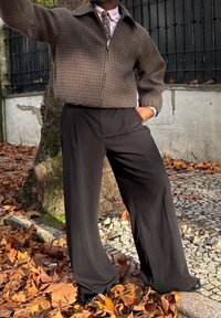 Veste zippée à motif pied-de-poule en marron et beige, associée à un pantalon noir ample. Scène avec des feuilles tombées et un pavé en pierre.