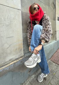 Egy piros kapucnis sálat, napszemüveget, szőrmével bélelt dzsekit, kék farmert és szürke sportcipőt viselő személy cipőt köt, miközben egy kőfalnak dől.