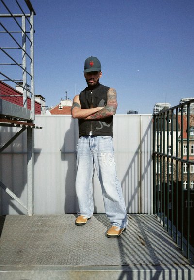 Chaqueta sin mangas negra, pantalones vaqueros anchos de color azul claro con detalle "KSW", calzado de color marrón claro, escenario urbano en la azotea con barandilla de metal y cielo despejado.