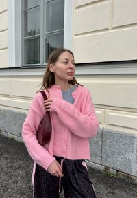 Pastelno roza pletena jakna z velikimi rokavi, z gumbom za zapiranje in rebrastim robom. V kombinaciji z hlačami z leopardjim vzorcem in burgundsko torbico.