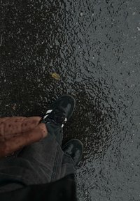 Mustat lenkkarit, joissa on valkoiset raidat märällä, tummalla asfaltilla. Tummanharmaat farkut ja ruskea takki näkyvät osittain kenkien päältä.