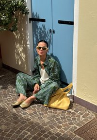 Zöld paisley mintás ruházat, krémszínű felső, sárga táska, arany cipők. Kék ajtón támaszkodva, köves úton ül, közelben növényekkel.