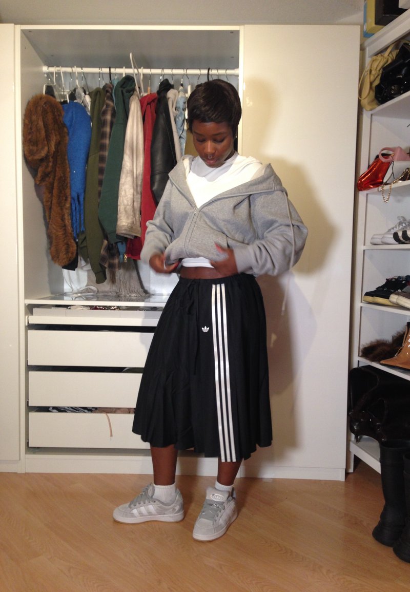 Una persona sistema una felpa grigia con zip sopra una maglietta bianca, indossando una gonna nera Adidas con strisce bianche e scarpe da ginnastica grigie in una stanza piena di vestiti e scarpe.