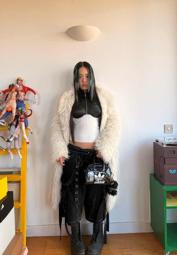Femme aux longs cheveux foncés portant un manteau en fausse fourrure blanc, un crop top noir, un pantalon large noir et des bottes noires, tenant un sac à main Adidas noir à l'intérieur.