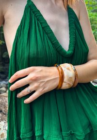 Robe verte plissée avec un décolleté plongeant ; main ornée de trois bracelets : ambre, beige marbré et texture bois. Bague étincelante visible.