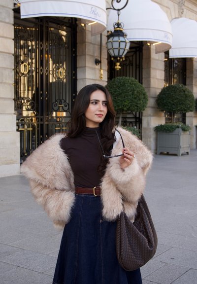 Mujer con abrigo de piel sosteniendo unas gafas de sol y un gran bolso tejido, de pie en una calle de la ciudad con árboles en macetas y entradas ornamentadas de edificios detrás de ella.