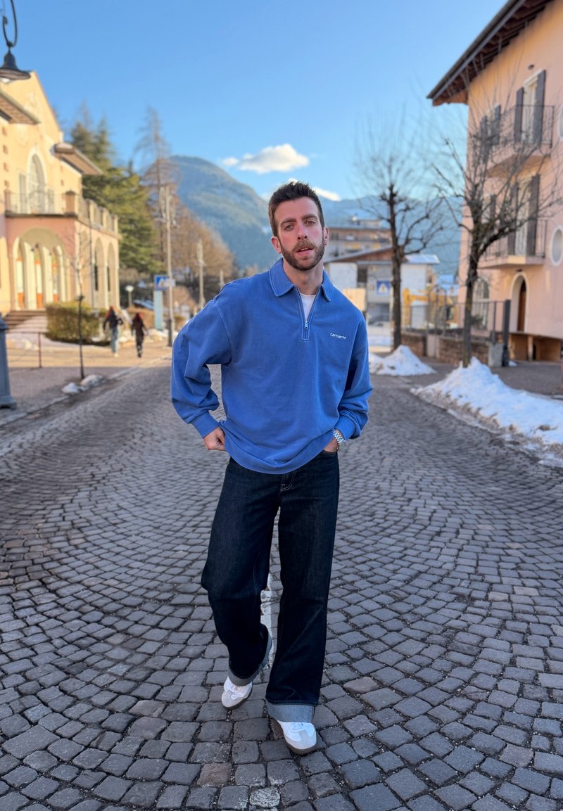 Άνδρας με μπλε πουλόβερ και σκουρόχρωμο τζιν που στέκεται σε δρόμο με λιθόστρωτο και χιονισμένα σημεία, με κτίρια και βουνά στο βάθος.