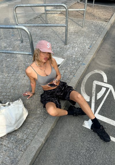 Modelo vistiendo un top corto gris, pantalones cortos de camuflaje negro y zapatillas negras. Accesorios incluyen una gorra rosa y collares en capas. Una bolsa de lienzo cercana.