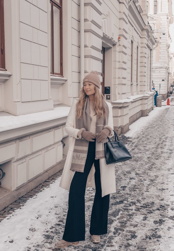 Frau trägt weißen Mantel, beige Hut, Schal und Handschuhe und steht auf einer verschneiten Straße, hält eine schwarze Handtasche vor einem Gebäude mit weißer Fassade.