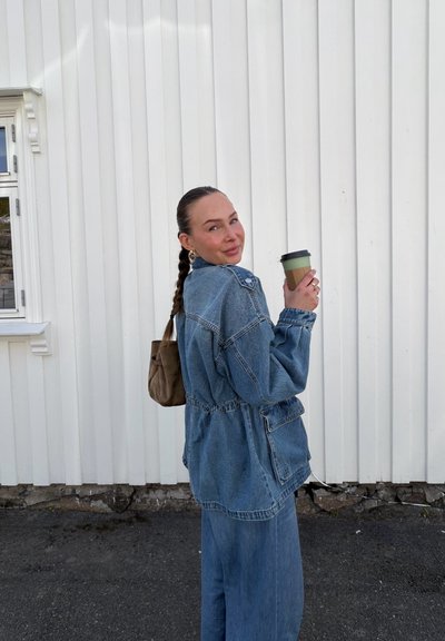Mujer con cabello trenzado lleva una chaqueta vaquera y pantalones holgados, sostiene una taza de café, de pie junto a una pared blanca con paneles.