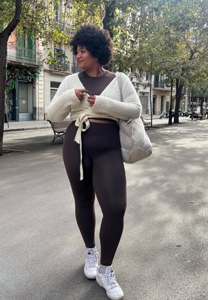 Conjunto de ropa deportiva marrón con un cárdigan corto atado en crema. Zapatillas blancas y una bolsa ligera completan el atuendo. Entorno urbano al aire libre con árboles.