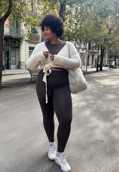 Conjunto de ropa deportiva marrón con un cárdigan corto atado en crema. Zapatillas blancas y una bolsa ligera completan el atuendo. Entorno urbano al aire libre con árboles.