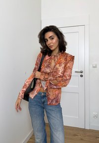 Ung kvinde med mørkt hår iført en gennemsigtig blomster-oberst i orange, blå jeans og sort skuldertaske, stående indendørs nær en hvid dør.