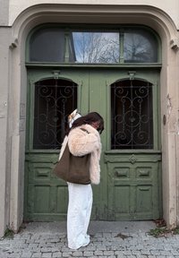 Brązowa pleciona torba na ramię z futrzanym kapturem płaszcza, stojąca przy zielonych drewnianych drzwiach z ozdobnymi żelaznymi kratami i kamiennej nawierzchni.