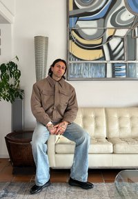 Veste en polaire marron avec grandes poches, associée à un jean évasé bleu clair et des chaussures noires, assis sur un canapé en cuir crème.