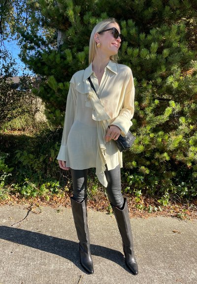 Blusa de seda crema con parte delantera con volantes, combinada con leggins de cuero negros y botas negras hasta la rodilla. Bolso negro llevado en bandolera.