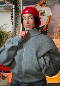 Siva jopa z zadrgo in visokim ovratnikom, velikimi rokavi in mehko teksturo, kombinirana z rde čo kapo. V ozadju so rastline in škatle za superge.