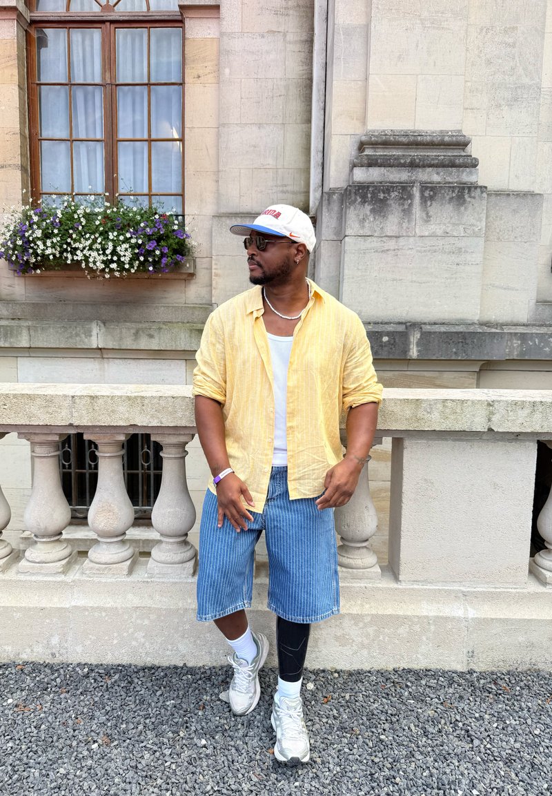 Homme portant une chemise jaune, un short bleu à rayures et une casquette blanche, debout sur du gravier près d'une balustrade en pierre avec des fleurs et une fenêtre derrière lui.