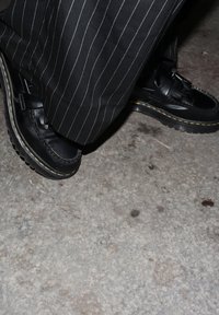 Mocasini negri din piele cu detaliu de cusătură și cataramă, asortați cu pantaloni negri cu dungi subțiri, pe o suprafață texturată de beton.