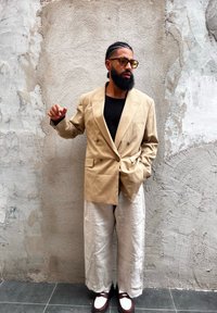 Blazer beige a doppiopetto, camicia nera, pantaloni ampi in lino chiaro e scarpe marroni con dettagli bianchi davanti a un muro grigio testurizzato.