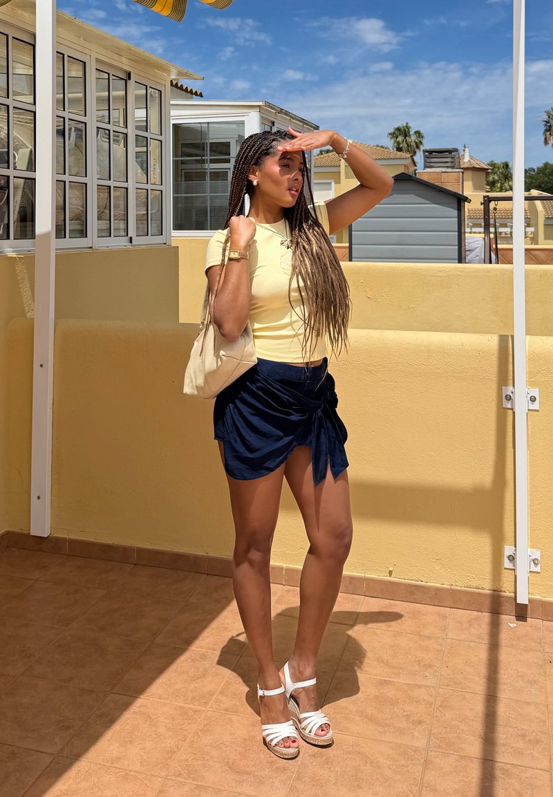 Femme aux longues tresses protégeant ses yeux du soleil, portant un haut jaune, une jupe marine, des sandales blanches et tenant un sac à main beige sur une terrasse ensoleillée.