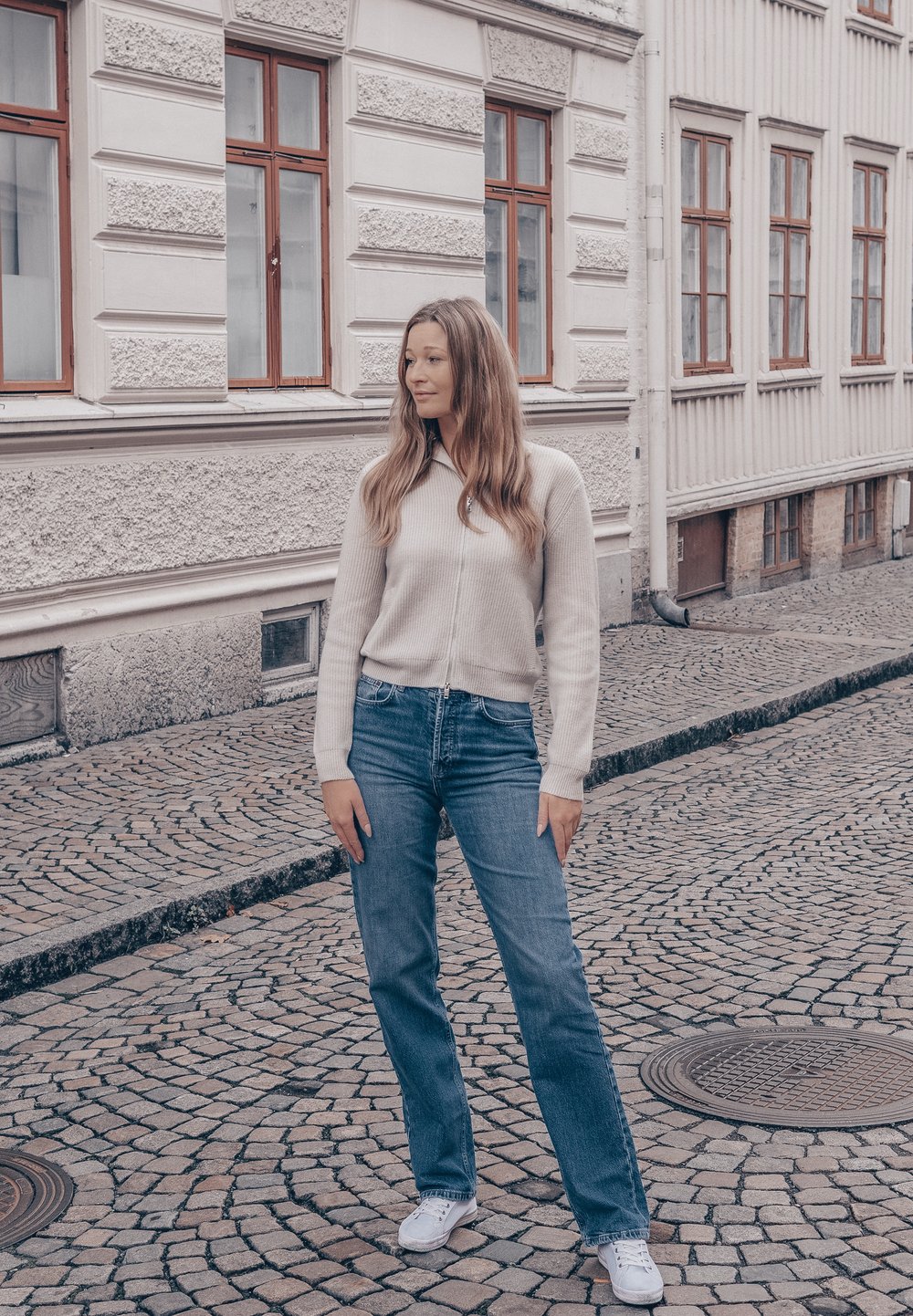 Lys beige genser med glidelås og ribbestruktur, blå denimjeans og hvite joggesko. Står på en brosteinsgate med mursteinsbygninger i bakgrunnen.