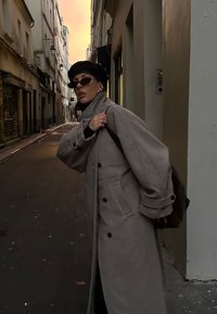 Sivý nadmerný kabát s veľkými gombíkmi, vysokým golierom a vreckami. Čierna pletená čiapka a slnečné okuliare. Hnedá taška nosená cez rameno na úzkej uličke.