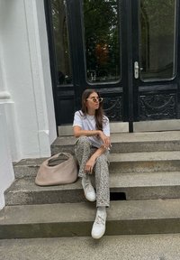 Béžová tkaná kabelka, bílý tričko a vzorované šedé kalhoty. Na sobě má světlé sluneční brýle a bílé tenisky. Sedí na kamenných schodech.