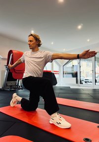 Fehér sportcipők piros és fekete részletekkel, fekete nadrág és fehér póló. A személy kitörést végez egy piros tornaszőnyegen.