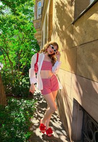 Czerwony w kratkę crop top oraz szorty w komplecie z białą, oversize'ową koszulą, czerwonymi sandałami z kwiatowym motywem i białymi okularami przeciwsłonecznymi na zielonym tle.