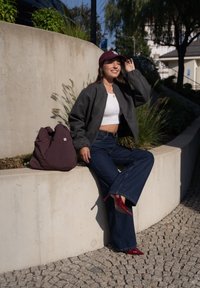Casaco bomber cinza, top cropped branco, jeans flared escuros, sapatos de salto altos vermelhos. Bolsa bordeaux encostada a uma parede de concreto em um ambiente ao ar livre.