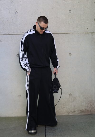 Chándal atlético negro con franjas laterales blancas, pantalones de corte holgado y chaqueta con cremallera. Sosteniendo una pequeña bolsa negra, de pie sobre una superficie gris.