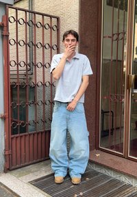 Camisa polo de manga curta cinza claro, jeans oversized azul claro, calçado castanho. Em pé à frente de uma parede padrão e de uma entrada com portão.